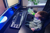 Happy baby is sitting at the computer keyboard with a bottle of milk in his hands. A child with a desktop PC in a home living room drinks milk formula from a bottle. Kid aged one year nine months