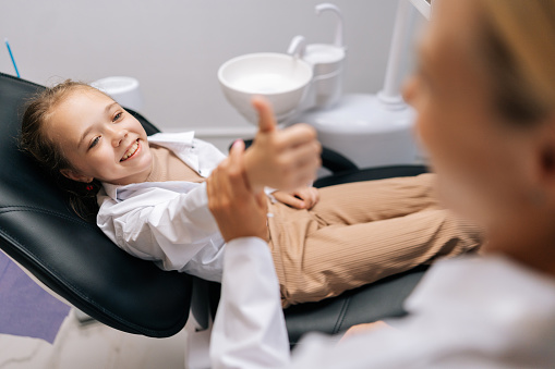 Top view of happy adorable little child girl patient showing thumbs up gesture to dentist because she liked pediatric dental service in modern dentistry clinic. Concept of children teeth treatment