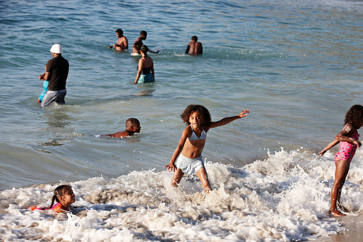 Cape Town, South Africa - January 8, 2023: The narrow beach at St James on the False Bay coastline is crowded with people during the vacation season, with children and adults splashing in the waves and women in the foreground beside a blue sun tent.