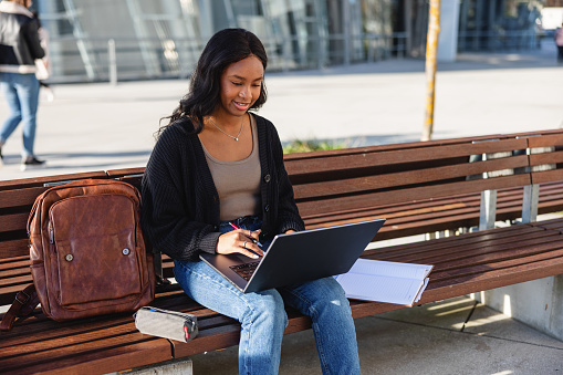 Three quarter shot with blurred background of a dark-skinned young Latin-American college student using her laptop a resource to study for an important exam while she is seated next to her brown backpack and notebook on a sunny day.