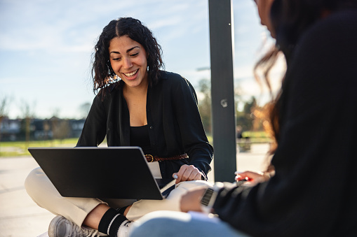 Three-quarter shot with blurred background of a young and beautiful Latin-American university student sitting with legs crossed on a wooden bench and carrying a laptop while talking to her classmate.