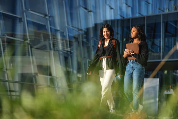 Multicultural College Students Walking On The University Campus Full length side shot with blurred background of two multicultural college students walking across campus while carrying their backpacks and notebooks for the next class on a sunny day, surrounded by grass and standing in front of a large glass-enclosed building. walking into glass door stock pictures, royalty-free photos & images