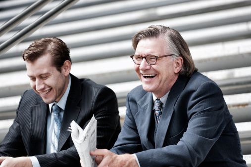 A young German businessman sitting on the exterior steps of a large office building, laughing with a more senior colleague