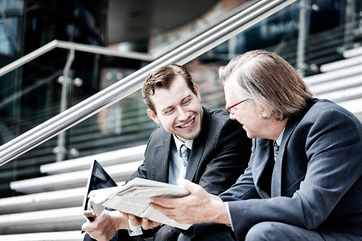 A young German businessman sitting on the exterior steps of a large office building, laughing with a more senior colleague