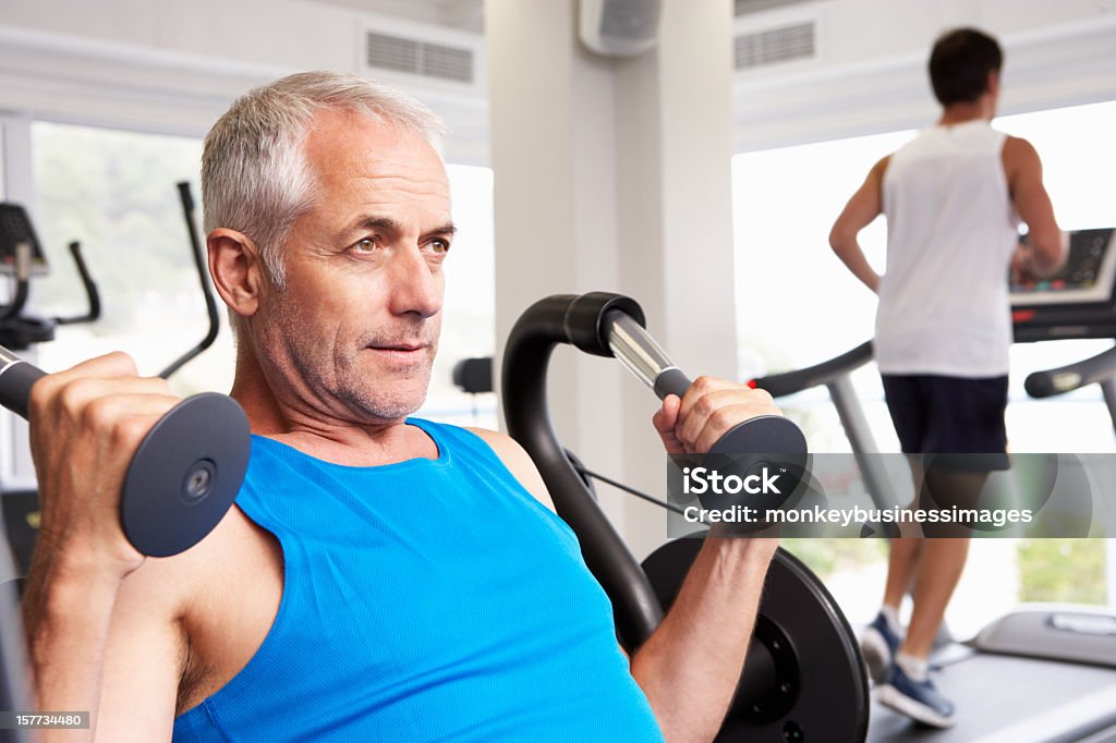 Man Using Weights Machine With Runner On Treadmill In Background Mature Men Stock Photo