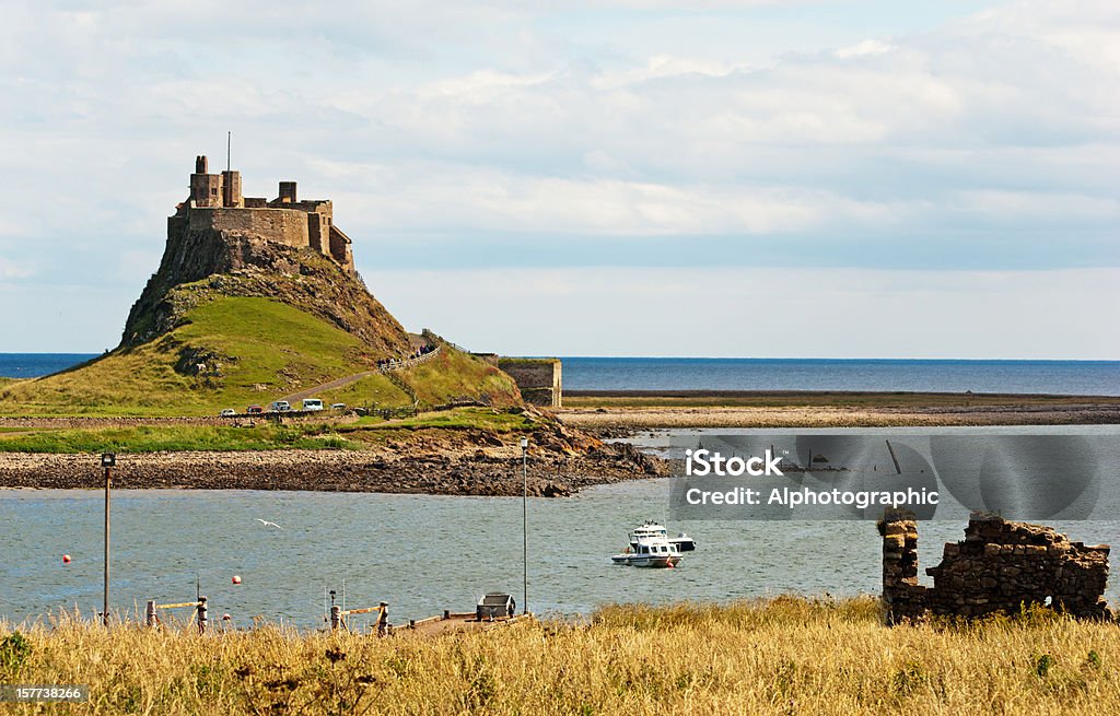 Lindisfarne Castle A view of Lindisfarne Castle on the Holy Isle in Northumberland, England against a pale summer sky with headland and boats in the foreground. Lindisfarne Castle Stock Photo