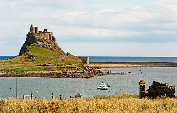 Lindisfarne Castle A view of Lindisfarne Castle on the Holy Isle in Northumberland, England against a pale summer sky with headland and boats in the foreground. farne islands stock pictures, royalty-free photos & images