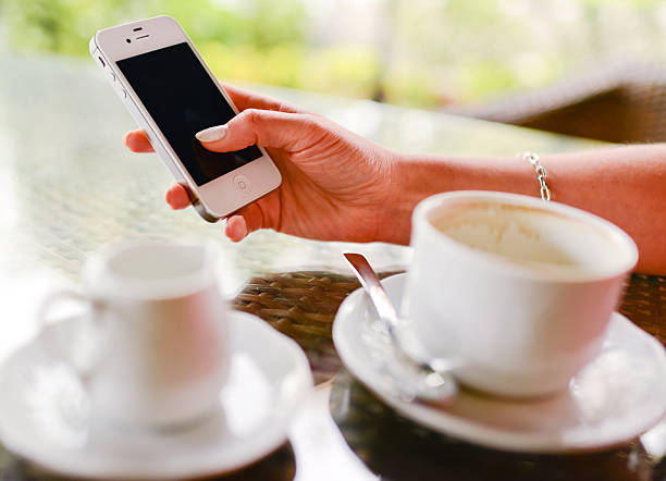 Man holding cellphone in his hand beside a cup of hot cappuccino coffee on the white table