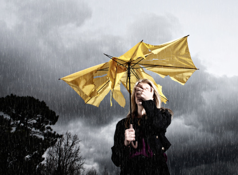 A blonde woman caught in a storm with a frayed and broken umbrella covers her face in nervous frustration; the thunderstorm has beaten her!