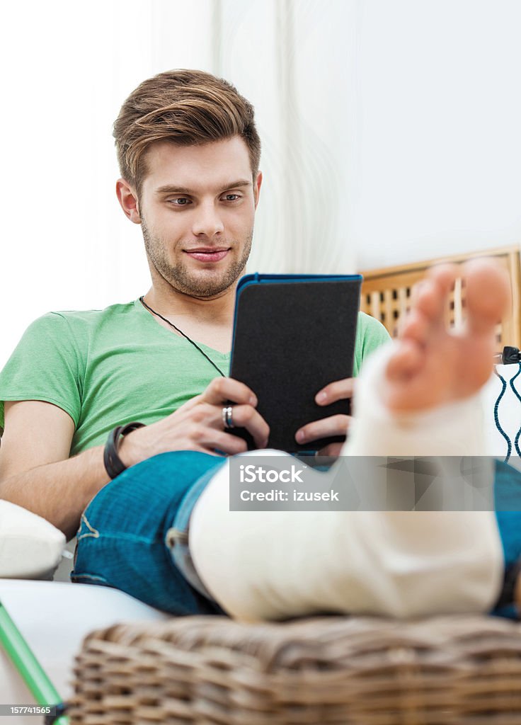 Young man with broken leg at home Young man with broken leg sitting on sofa at home and reading e-book. Focus on background. Home Interior Stock Photo