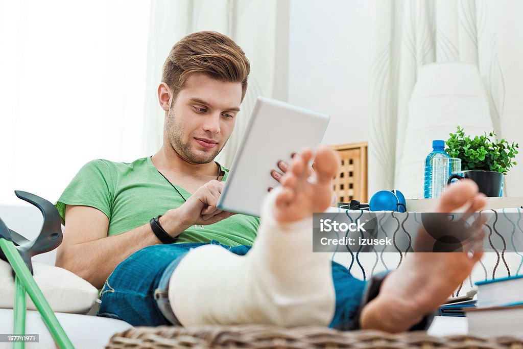 Young man with broken leg at home Young man with broken leg sitting on sofa at home and using digital tablet.  Orthopedic Cast Stock Photo