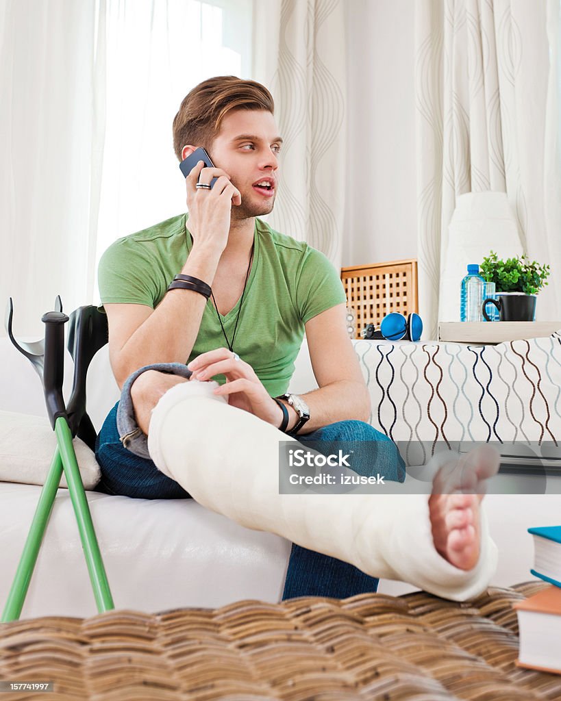 Young man with broken leg at home Young man with broken leg in plaster cast sitting on sofa at home and talking on the mobile phone.  Orthopedic Cast Stock Photo
