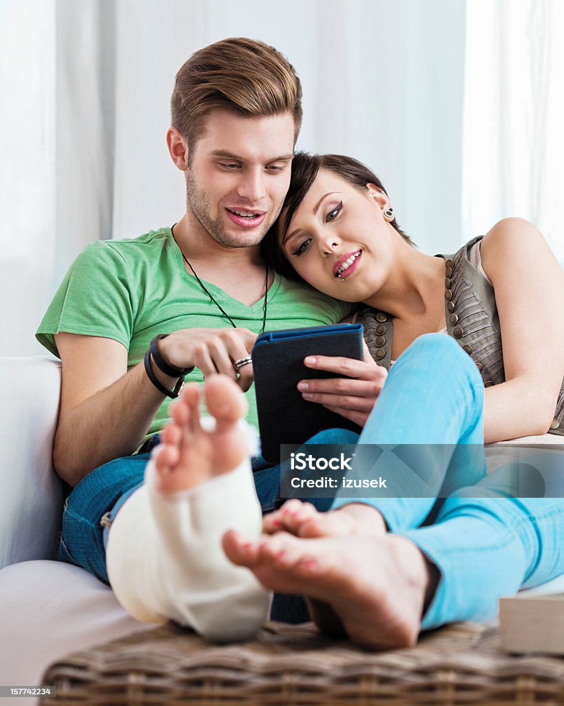 Young couple using e-reader Young couple sitting on sofa at home and using e-reader together. Man has broken leg in a plaster cast. Happiness Stock Photo