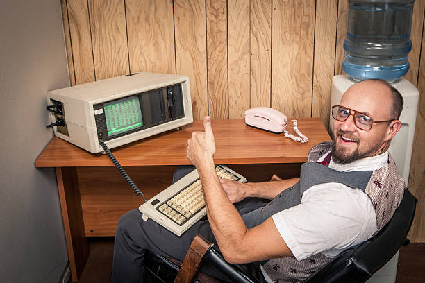 Thumbs up computer worker nerd  on phone at cubicle Happy office worker sitting, typing, working, and selling big at 1970's or 1980's monochrome computer.  Retro office interior scene.  Wood Paneling and a water cooler in the background. Old retro vintage technology uncool stock pictures, royalty-free photos & images