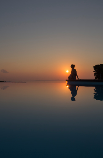 Young woman sitting by the edge of an infinity pool: her profile to the horizon over the sea, she meditates on the sunrise. Unrecognizible person-silhouette. Shot 1