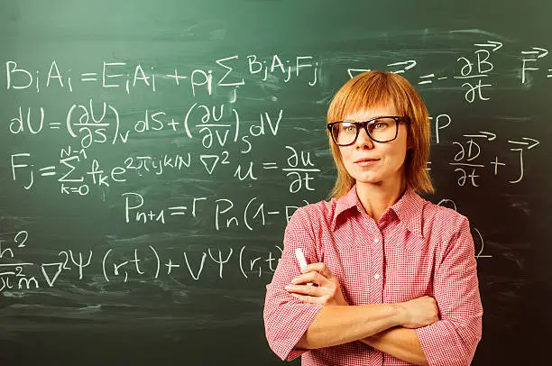 Close-up on female teacher in horn rimmed glasses. She is standing against chalkboard and holding chalk. Focus on blackboard.