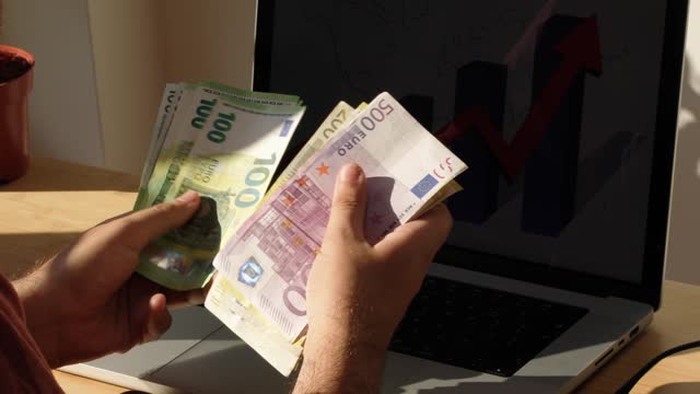 Unrecognizable man counting euro banknotes, while working on laptop