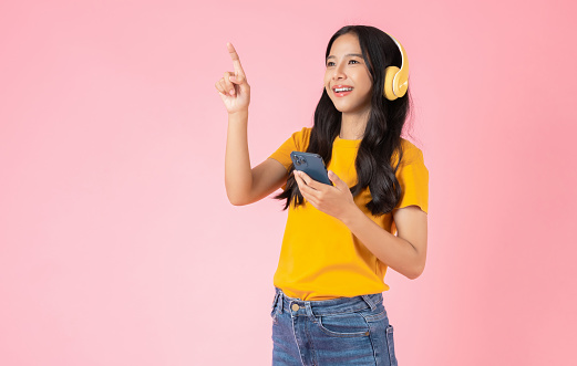 Cheerful young Asian woman in headphones listening to music and enjoy favourite playlist application on smartphone with shows hand pointing to the front on pink background.