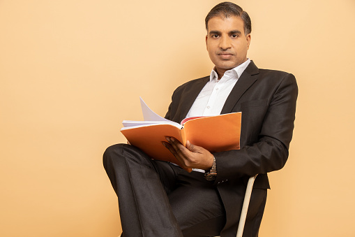 Mature indian man wearing black suit reading book while sitting on chair isolated over beige background.