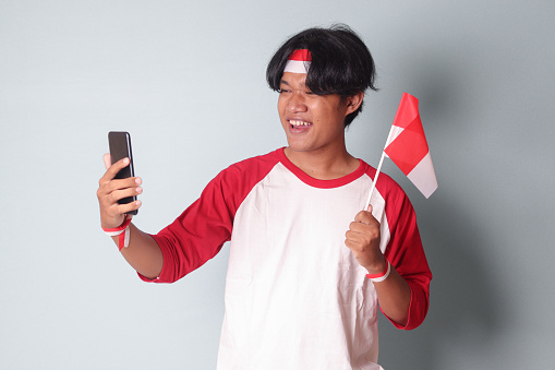 Portrait of attractive Asian man in t-shirt with red and white ribbon on head, taking a picture of himself while holding Indonesia flag. Isolated image on gray background