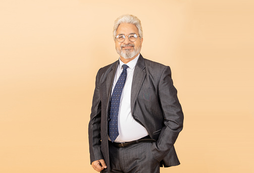 Happy senior indian businessman wearing black suit and tie standing isolated over beige background. Corporate concept