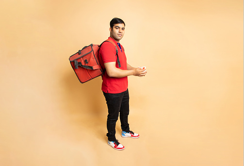Young indian delivery man with thermal insulated bag standing isolated over beige background. Food order and distribution service.