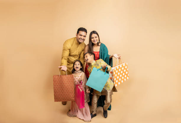쇼핑백을 들고 전통 의상을 입은 행복한 젊은 인도 가족과 함께 베이지색 스튜디오 배경에 고립된 디왈리 축제를 축하한다. 아이들과 함께 하는 부모들은 축제 시즌 세일을 축하합니다 - kurta 뉴스 사진 이미지