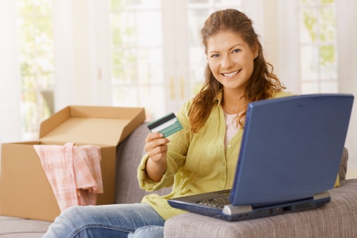 Happy young woman, sitting on sofa in living room, purchasing on laptop with credit card, smiling, received goods behind her...