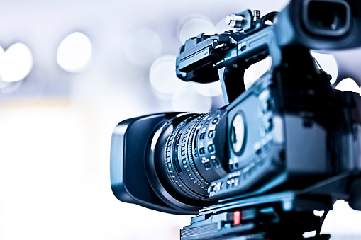 Female news reporter in front of camera, video camera is in the focus, reporter in the background is blurred, Nikon Z7