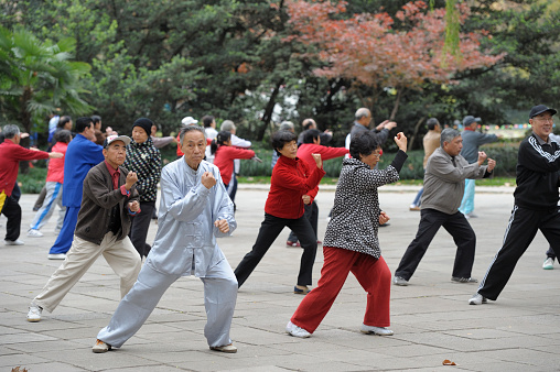 A small group of seniors stand outside together as they participate in a Tai Chi class. They are each wearing comfortable t-shirts and focused on their hand movements.