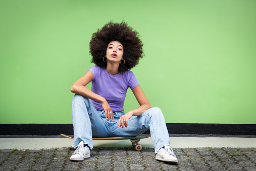 Full body of young African female in Afro hairstyle and casual clothes with makeup sitting on skateboard over terraced floor and looking at camera against green wall