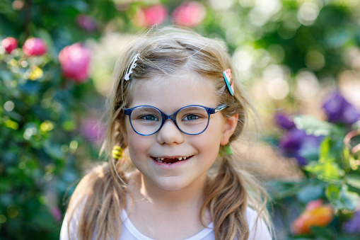 Portrait of a cute preschool girl with eye glasses and big teeth gap outdoors in park. Happy funny child on sunny summer day. Kid loosing milk tooth.