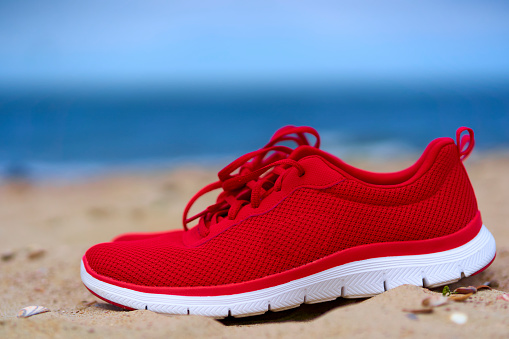 Red ladies sneaker shoes on empty sandy beach. Footwear for sport and leisure on the sea. Close up.