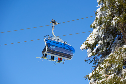 Feldberg, Germany - February 08, 2023: Leitner Ropeways against blue sky. Full gondola with winter sports enthusiasts. Fir tree with snow and icicles. Sunny weather. Low angle view. Black forest.