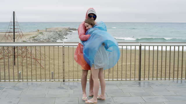 Couple holding each other hugging wearing multi colored raincoat poncho in rainy weather on the beach  in wind