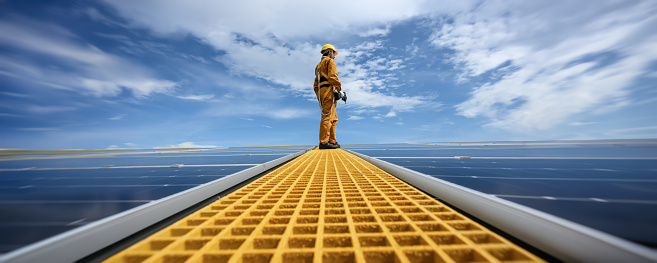 Technician Young Wearing Safety Protective Clothing on Walkway with Tool Standing to Checking Quality of Solar Panel or Photovoltaic Installation in Daytime on Factory Roof Buildings.