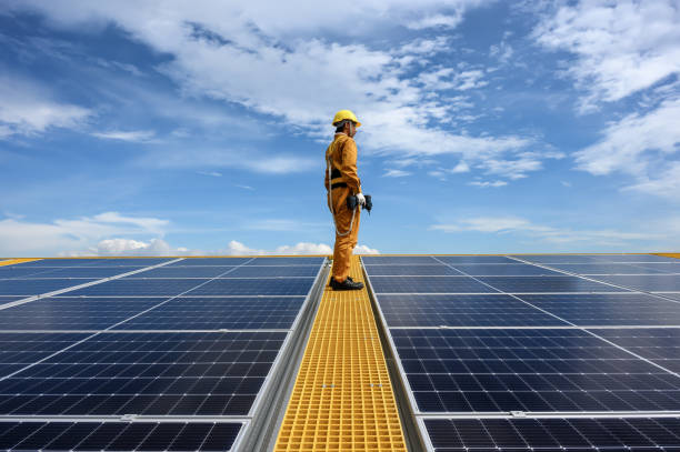 técnico joven que usa ropa de protección de seguridad en la pasarela con herramienta de pie para verificar la calidad del panel solar o la instalación fotovoltaica durante el día en los edificios del techo de la fábrica. - solar panel engineer solar power station solar energy fotografías e imágenes de stock