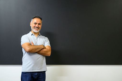 Happy smiling instructor with his arms crossed standing at the blackboard in a classroom