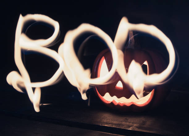 Word Boo drawn with light painting fire. Ceramic Halloween Jack-o'-lantern pumpkin with a candle in background.