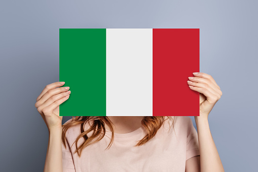 Student girl holds blank white speech bubble with italian flag isolated over grey studio background. woman holding a round information piece of paper dialog
