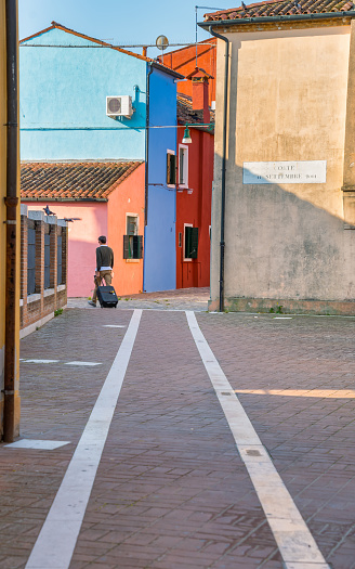 Venice, Italy - May 30 2023: Unidentifiable rear view of a solo traveler with a trolley bag on the narrow colorful alleys of Burano, Venice