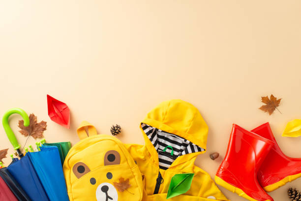 stylische regenbekleidung für kinder bei nassem wetter. top-down-bild mit bunten gummistiefeln, einem regenmantel, einem rucksack und einem regenschirm auf einem beigefarbenen isolierten hintergrund, mit platz für text oder werbung - puddle rain boot water stock-fotos und bilder