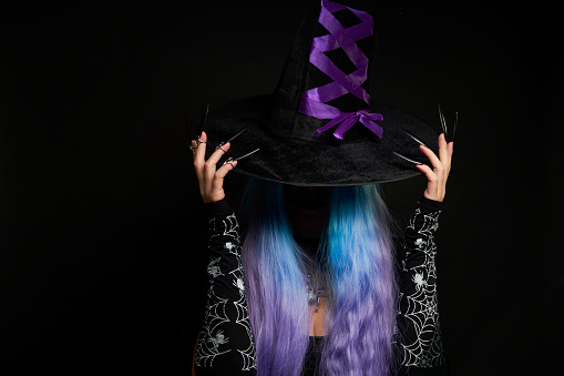 Halloween witch woman in black dress, cloak, black hat with violet ribbon, blue and violet hair and long black nails. Halloween celebration concept