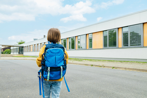 A view from the back of a girl with a school bag on her back is standing next to the school building. Beginning of the school year.