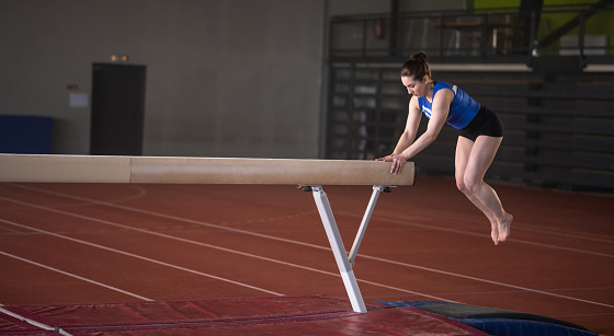 Female gymnast doing a complicated trick on balance beam in gym. Sport and endurance concept.