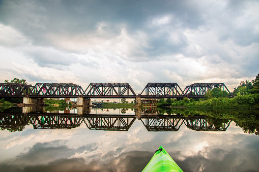 A multi segment trestle train track is going across the Scioto River in downtown Columbus Ohio.  There is construction happening on the right side of the bridge.  The scene is from the point of view (POV) of a kayaker on the river.  The scene is reflected in the river.