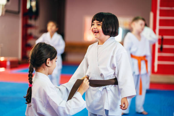 um grupo de crianças do ensino fundamental está tendo uma aula de artes marciais. amigos amarrando um cinto em um dojo - karate child judo belt - fotografias e filmes do acervo