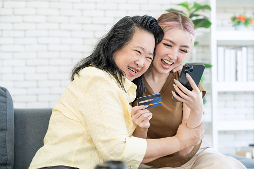 Happy Asian family, young daughter and senior elderly mom hugged each other using smartphone online shopping use credit card payment online application at living room at home.