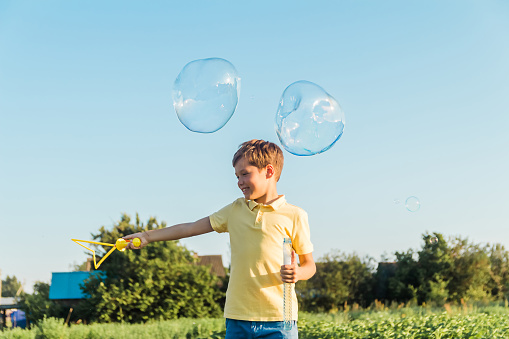 Caucasian boy launches big soap bubbles on a sunny summer day outdoors. Active leisure, happy childhood