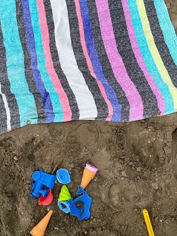 Stock photo showing close-up, elevated view of a collection of children's multicoloured, plastic beach toys on sandy beach besides a vibrant coloured striped towel. Toys include spades, sea animal moulds, ice cream cones and a steam engine.